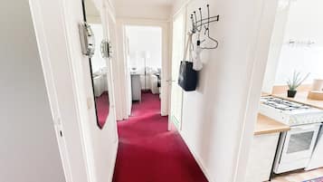 A hallway with a red carpet, white walls, a mirror, coat hooks, a small table with a lamp, and a kitchen area with a gas stove.