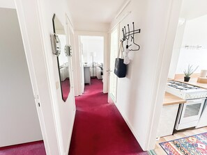 A hallway with a red carpet, white walls, a mirror, coat hooks, a small table with a lamp, and a kitchen area with a gas stove. - Vacation Rental Behind the Church in Döse (Cuxhaven)