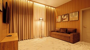 A room with a wooden panel wall, a brown sofa, a standing floor lamp, and a flat-screen TV on a wooden side table. - Europa  (Semey)