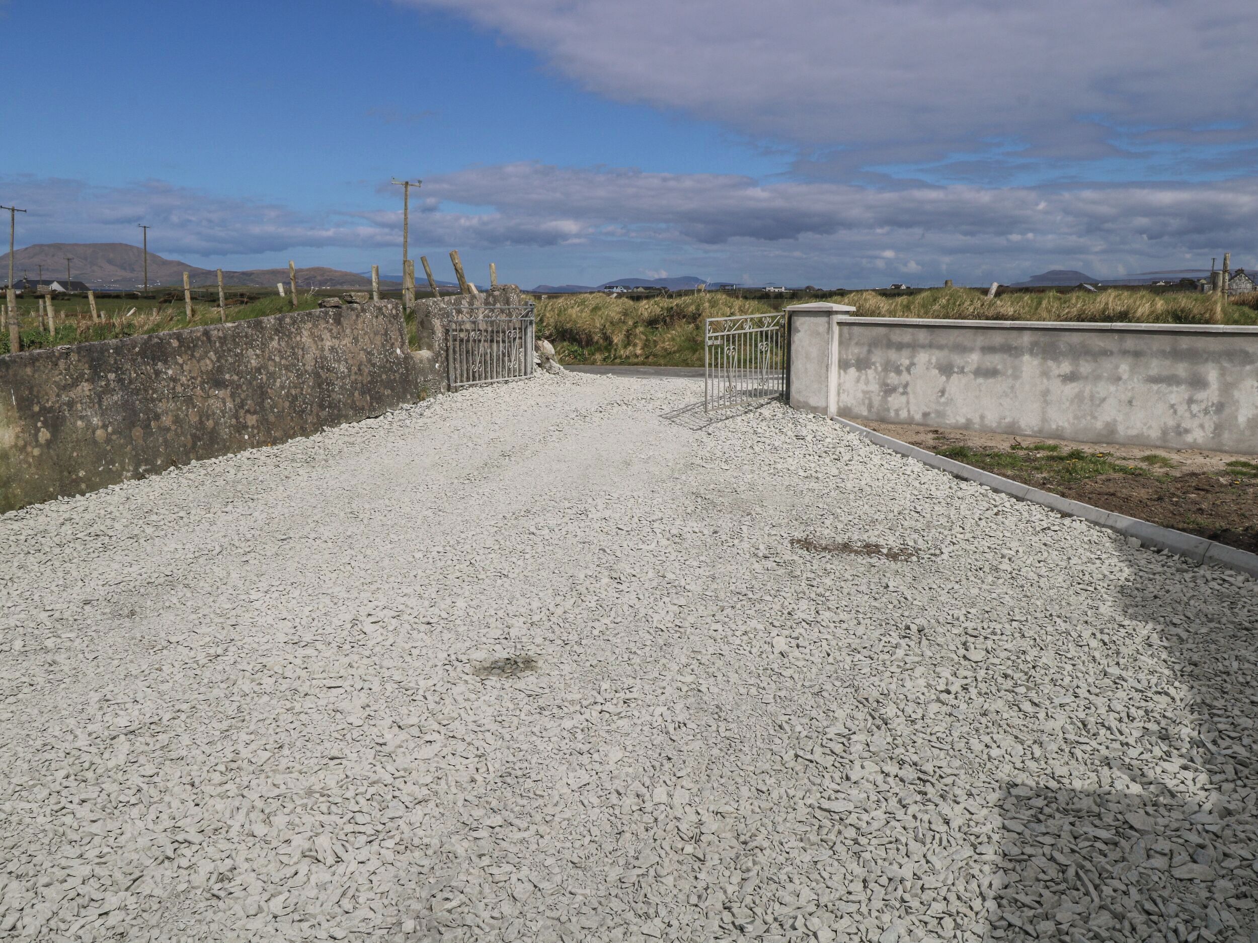 Een grindpad dat leidt naar een toegangspoort met een betonnen muur en een metalen hek.