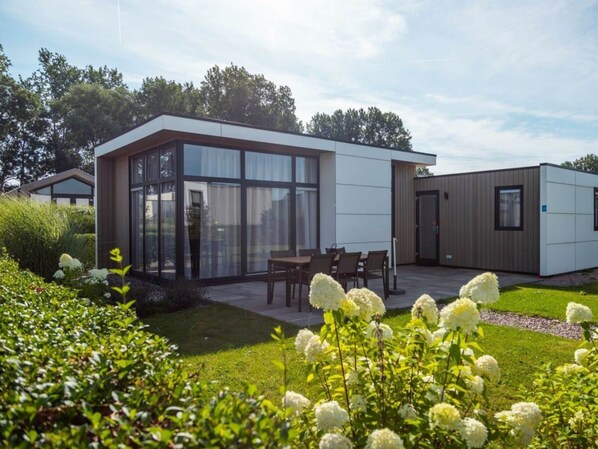 Ein modernes Gebäude mit großen Fenstern, einer Terrasse mit Sitzgelegenheiten und einem Garten mit weißen Blüten.