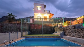 Una piscina con borde de piedra, una casa de dos pisos con balcón y un muro de piedra.