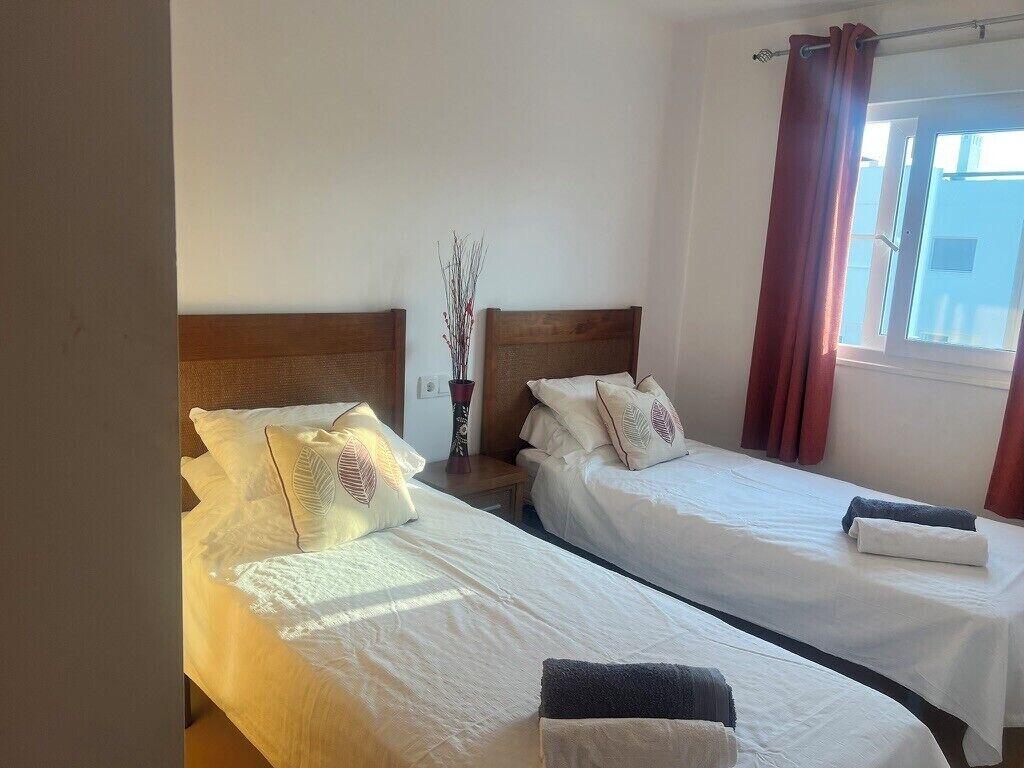 Habitación de hotel con dos camas, una mesita de noche, una ventana con cortinas y un florero con ramas.