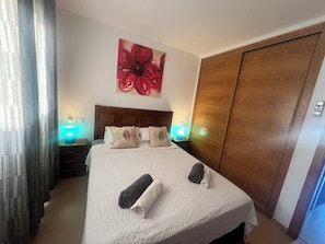 A hotel room with a bed, two bedside lamps, a wooden wardrobe, and a large floral artwork on the wall.