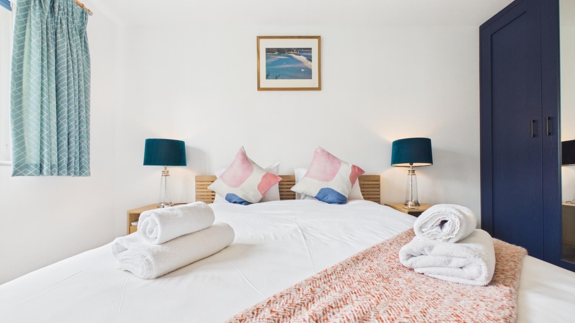 A hotel room with a bed, two bedside tables with lamps, a blue wardrobe, and a framed picture on the wall.
