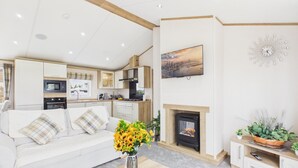 A modern living room with a sofa, a fireplace, a kitchen area, and a painting on the wall. - Kittiwake Lodge - 2 Bed Lodge - Wisemans Bridge (Narberth)