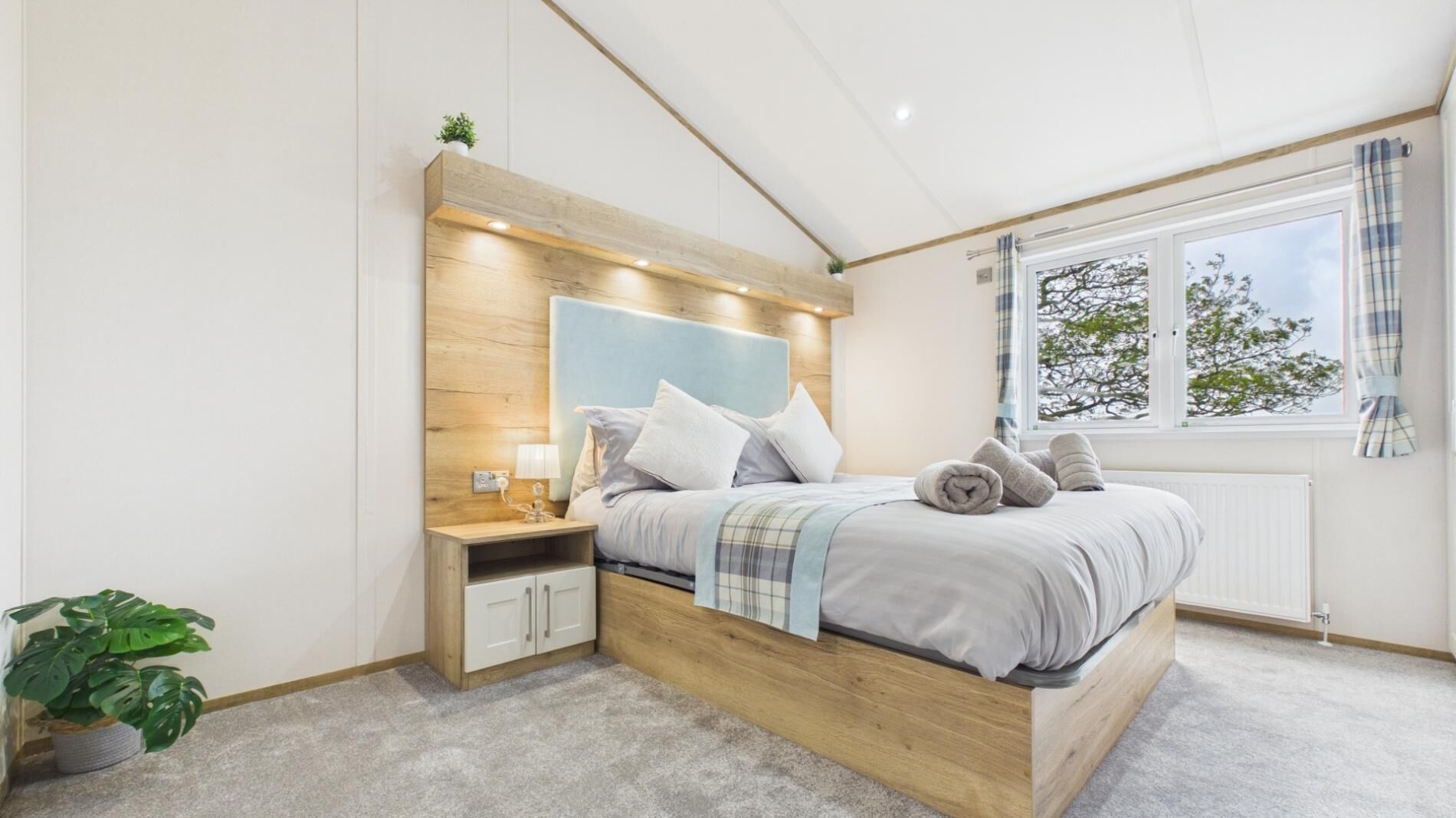 Camera da letto con testiera in legno, comodino con lampada, finestra con tende e una pianta in vaso.