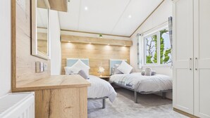 A bedroom with two beds, a wooden headboard, a bedside table, a radiator, and a window with a view of trees. - Kittiwake Lodge - 2 Bed Lodge - Wisemans Bridge (Narberth)