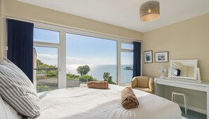 Ein Hotelzimmer mit einem großen Bett, einem Sessel, einem Schreibtisch mit Spiegel und Meerblick.