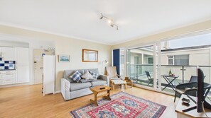 Ein Wohnzimmer mit Sofa, Sessel, einem kleinen Tisch, einem gemusterten Teppich und einem Balkon mit Gartenmöbeln.