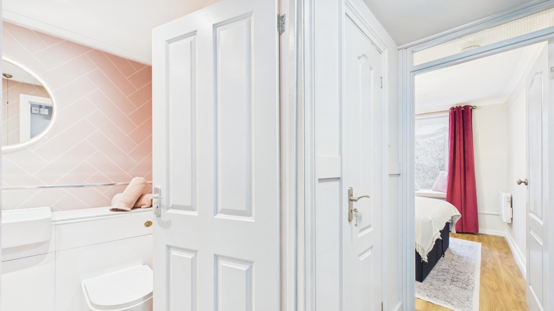 A bedroom with a bed, a window with curtains, a bathroom with a mirror, and a door.