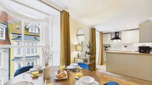 A dining area with a table set for breakfast, a kitchen with modern appliances, and a view of traditional buildings through large windows. - Harbour Rose - 1 Bedroom Apartment - Tenby (Tenby)
