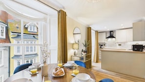 Un comedor con una mesa dispuesta para el desayuno, una cocina con electrodomésticos modernos y vistas a edificios tradicionales a través de amplias ventanas.