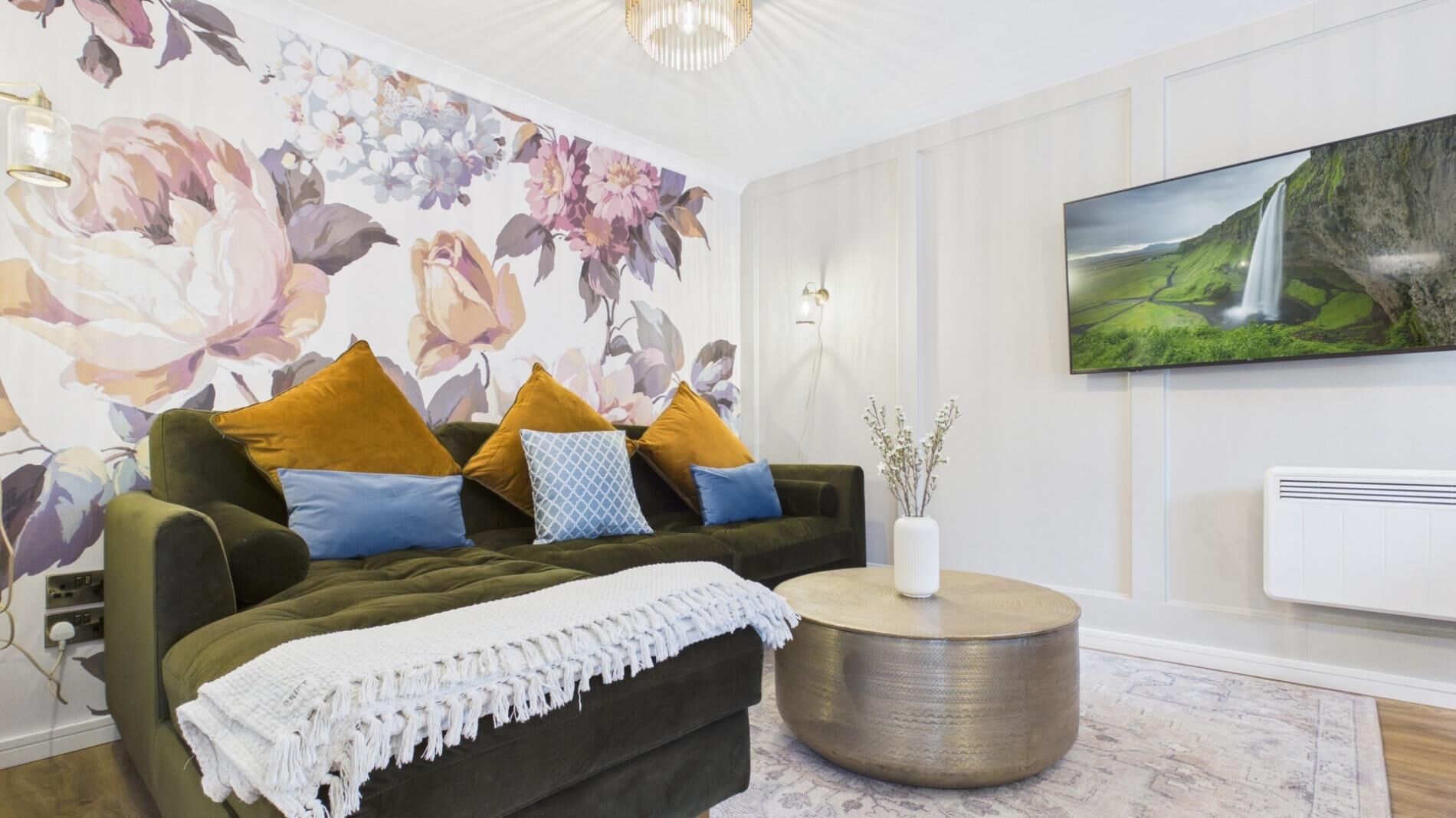 A living room with a green sofa, mustard and blue cushions, a white throw blanket, a round side table with a vase, and a large floral wallpaper.