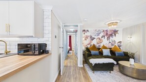 Eine moderne Küche mit Mikrowelle, einer Holz Arbeitsplatte und einem goldenen Wasserhahn. Ein Flur führt zu einem Wohnbereich mit einer Couch und einem runden Couchtisch.