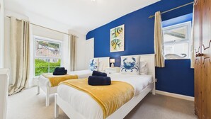 A bedroom with two beds, a window with curtains, a blue wall, and a view of houses outside. - St Catherines Apartment - 2 Bedroom - Penally (Tenby)