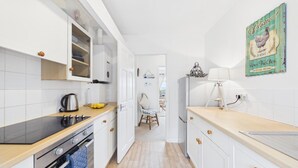 A narrow kitchen with white cabinets, a stove, and a chair in the background. - St Catherines Apartment - 2 Bedroom - Penally (Tenby)