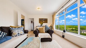 A living room with a sofa, a coffee table, and a view of a green landscape through large windows. - St Catherines Apartment - 2 Bedroom - Penally (Tenby)