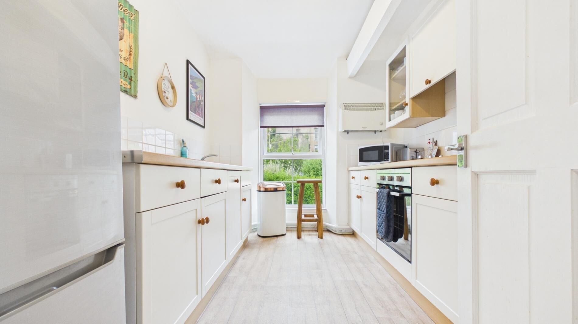 A narrow kitchen with white cabinets, a microwave, an oven, and a window with a view of greenery.