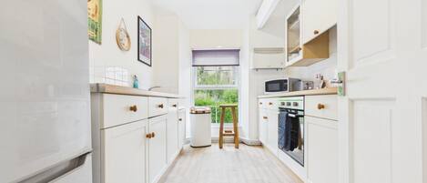 A narrow kitchen with white cabinets, a microwave, an oven, and a window with a view of greenery.