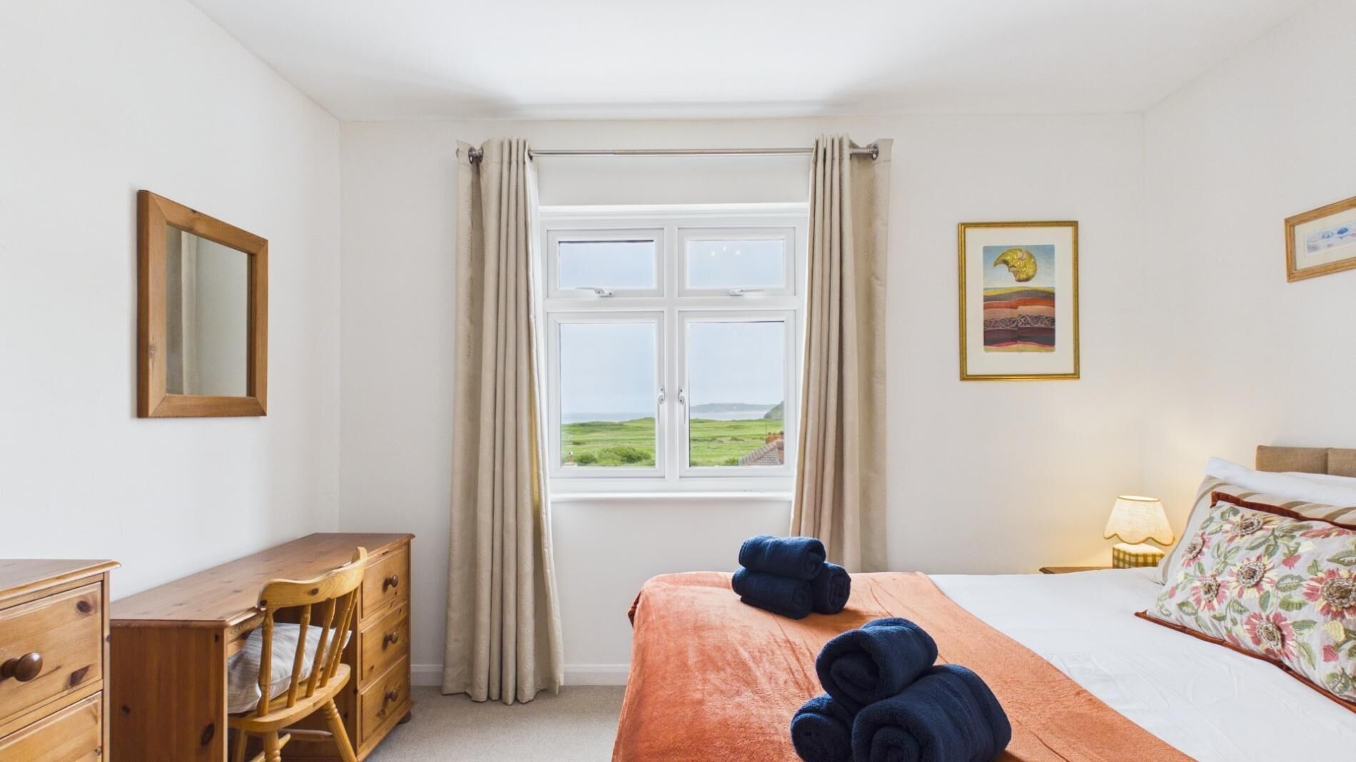 A hotel room with a bed, a desk, a chair, a window with curtains, and a painting on the wall.