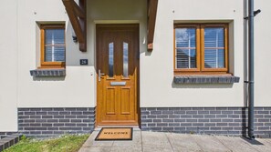 Ein Haus mit Holztür, zwei Fenster mit Fensterläden und einem Schild mit der Aufschrift '28'. Vor der Tür liegt eine Fußmatte mit dem Schriftzug 'WILLKOMMEN'.