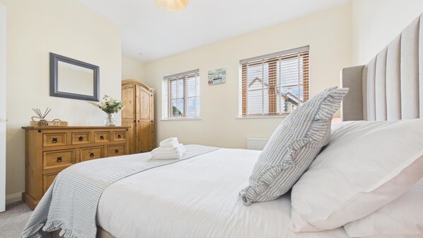A bedroom with a bed, a wooden dresser, a mirror, and two windows with shutters. - Anchors Aweigh - 2 Bedroom - New Hedges (Saundersfoot)