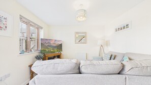 Un salon avec un canapé gris, un téléviseur à écran plat posé sur un meuble en bois, une fenêtre avec des stores, un radiateur et un luminaire au plafond.