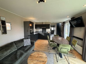 A modern living room with a sofa, a dining table with chairs, a kitchen area, and a TV mounted on the wall. - Pavilion L"etage 6 (Kaatsheuvel)