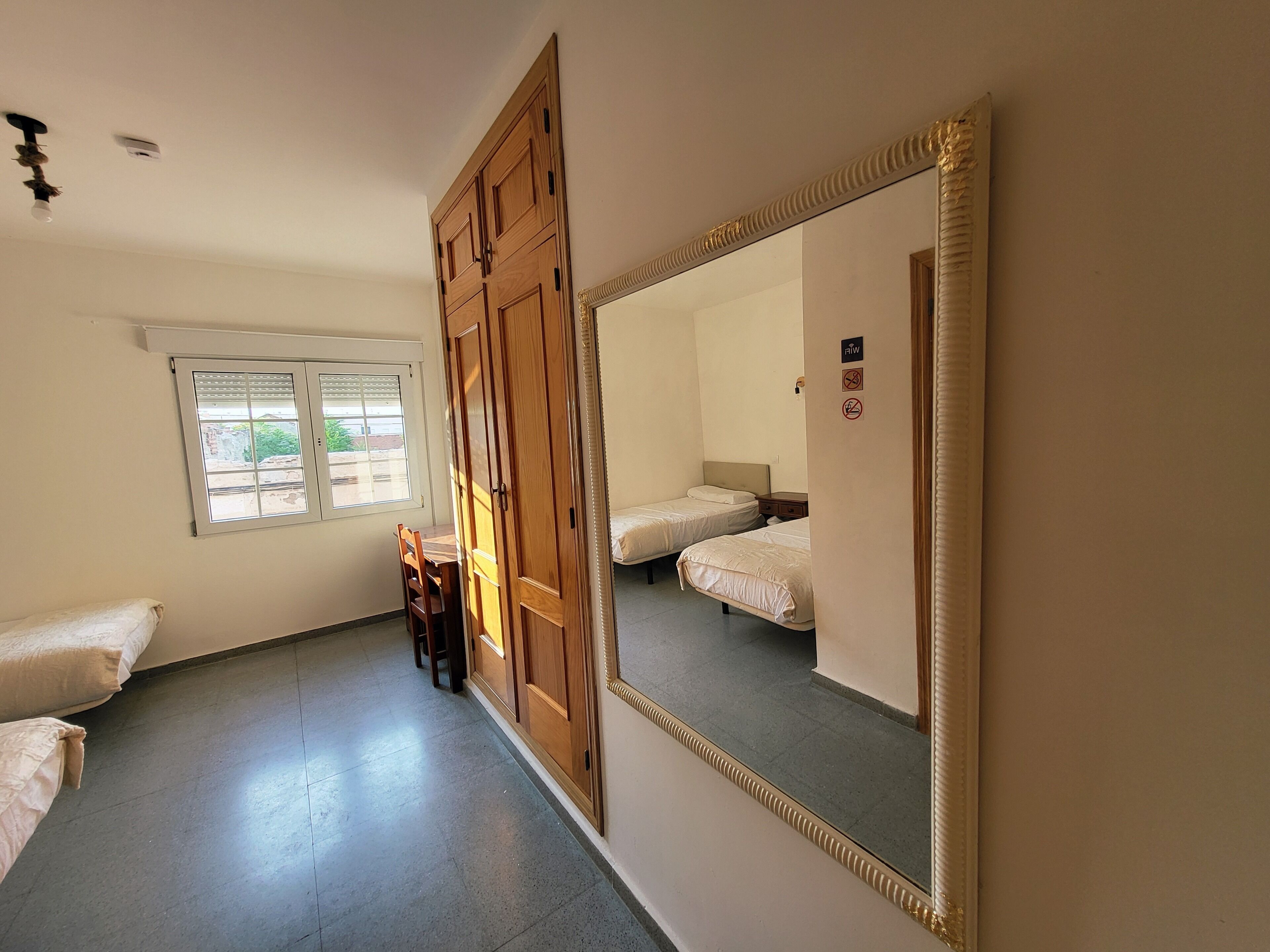 Ein Zimmer mit zwei Betten, einem Holzschrank, einem Fenster und einem Spiegel.