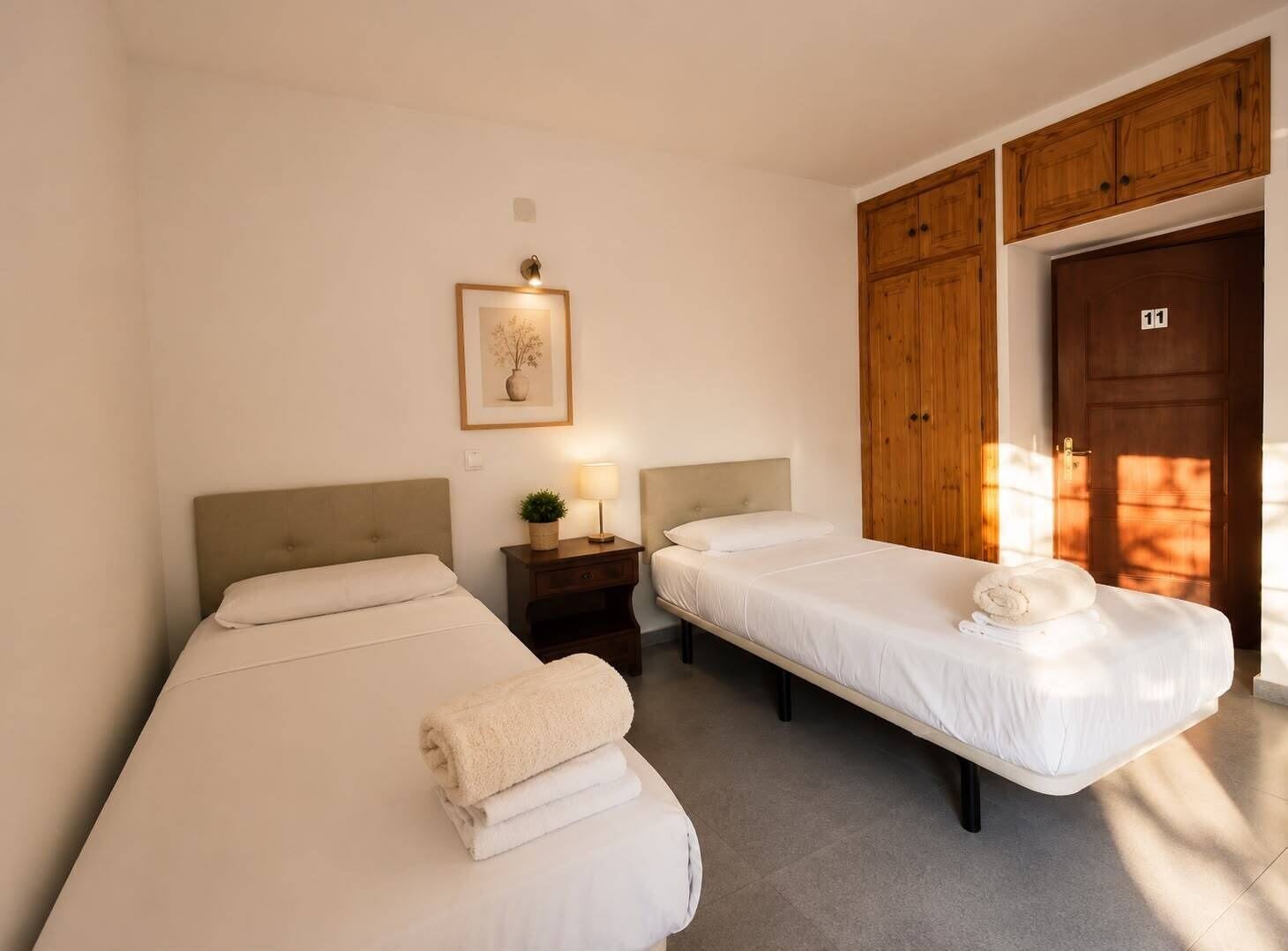 Habitación de hotel con dos camas, una mesita de noche con lámpara, un cuadro enmarcado en la pared y armarios de madera.