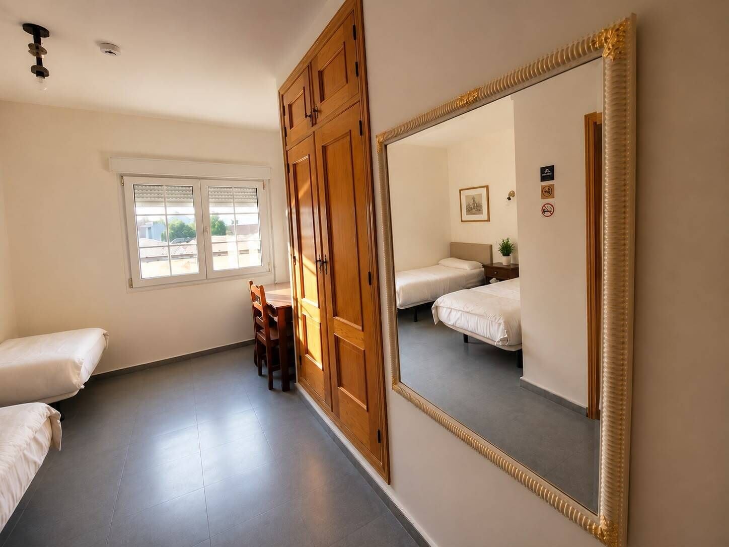 A hotel room with two beds, a wooden wardrobe, a large mirror, a window with blinds, and a small table with a plant.