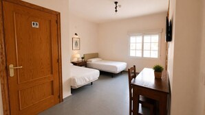 A room with two single beds, a wooden door, a desk with a chair, a small table with a plant, a wall-mounted TV, and a window with blinds.