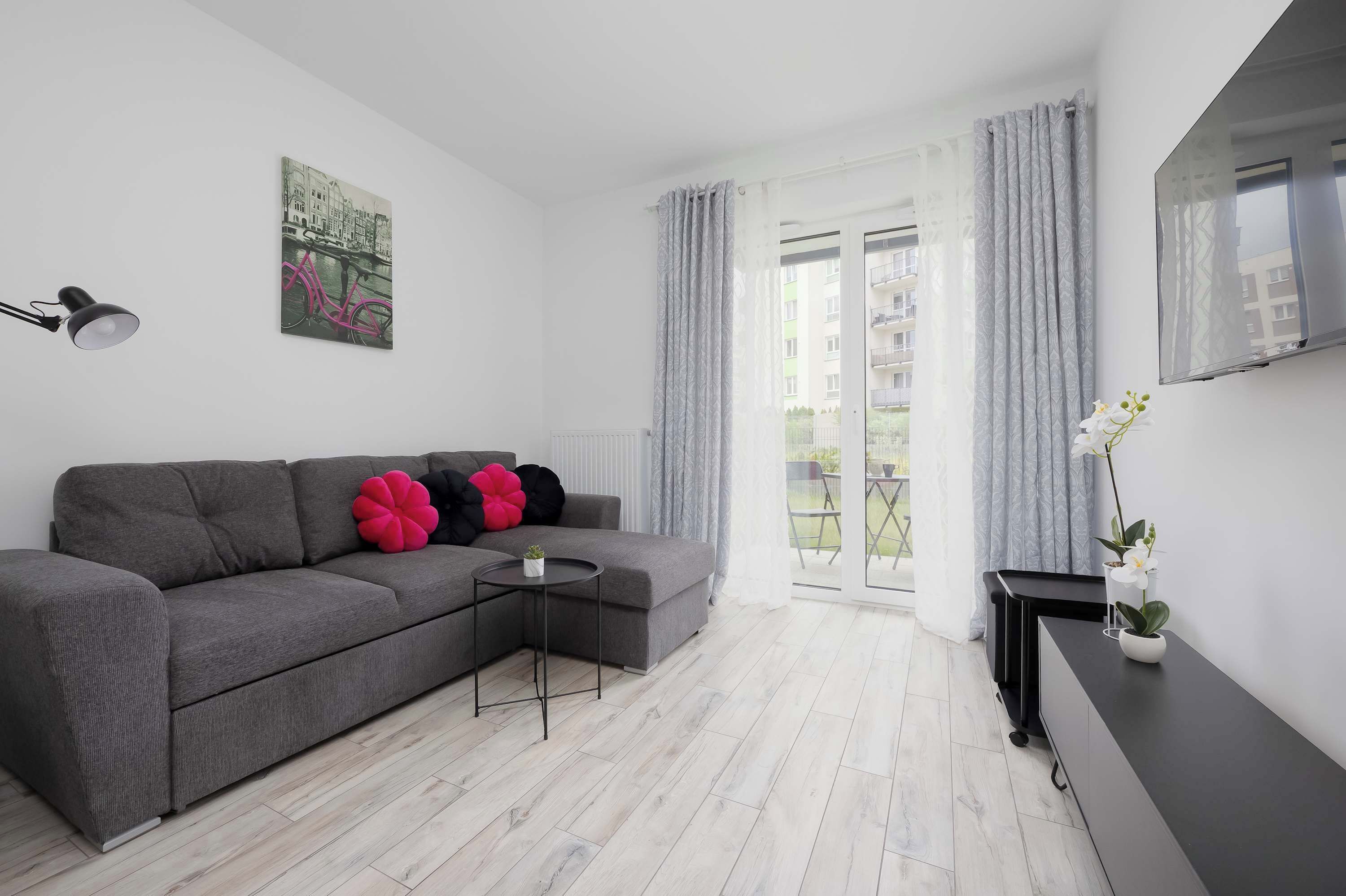 A modern living room with a grey sofa, a small side table, a flat-screen TV, and a balcony with a view.