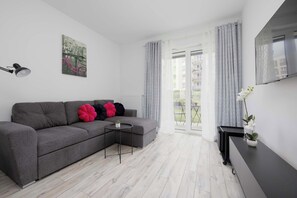 A modern living room with a grey sofa, a small side table, a flat-screen TV, and a balcony with a view. - Stylish Apartment with Garden | Quiet Neighborhood (Warszawa)