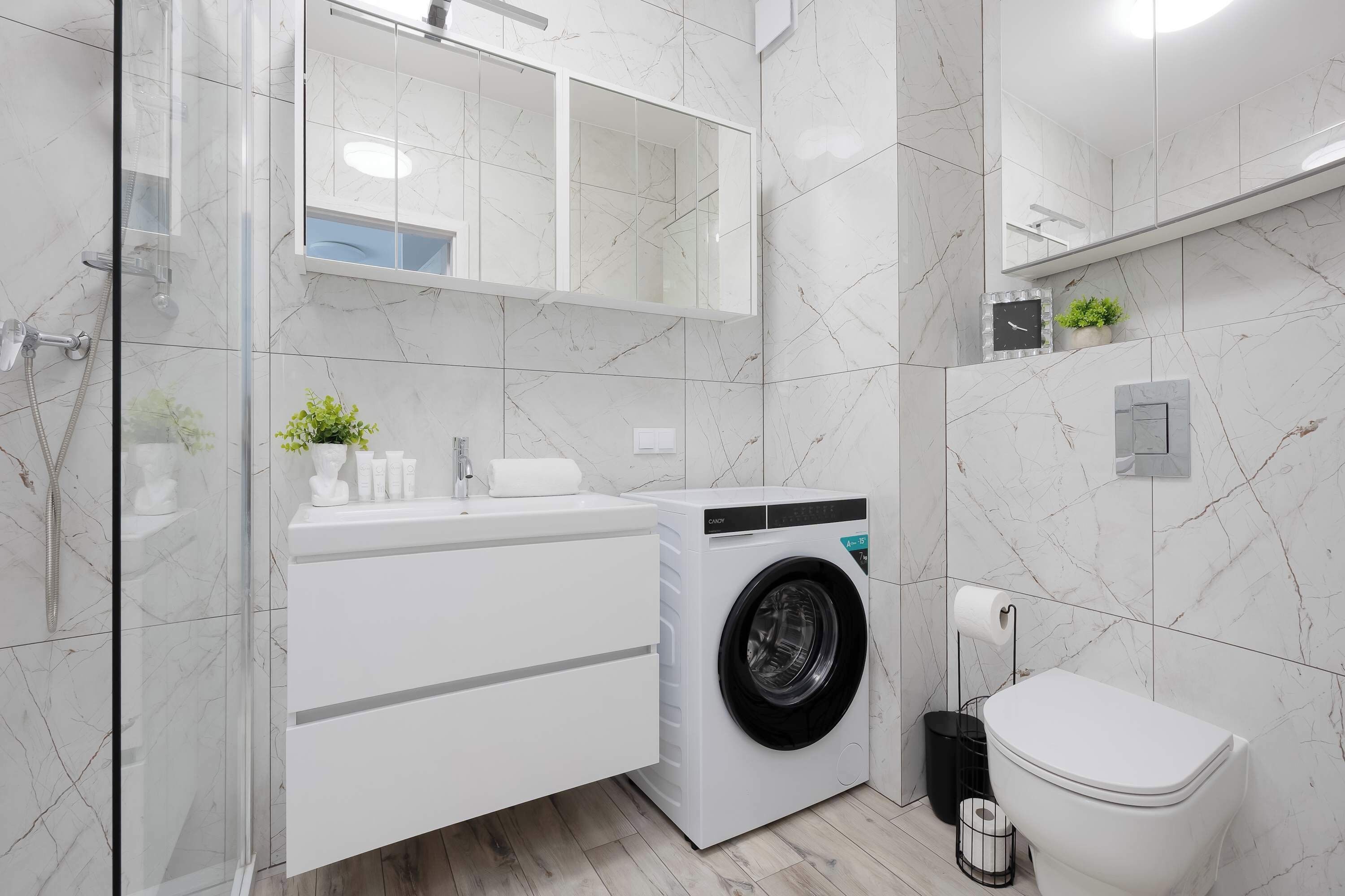 A modern bathroom with a glass shower enclosure, a built-in washing machine, a toilet, and a mirror above the sink.