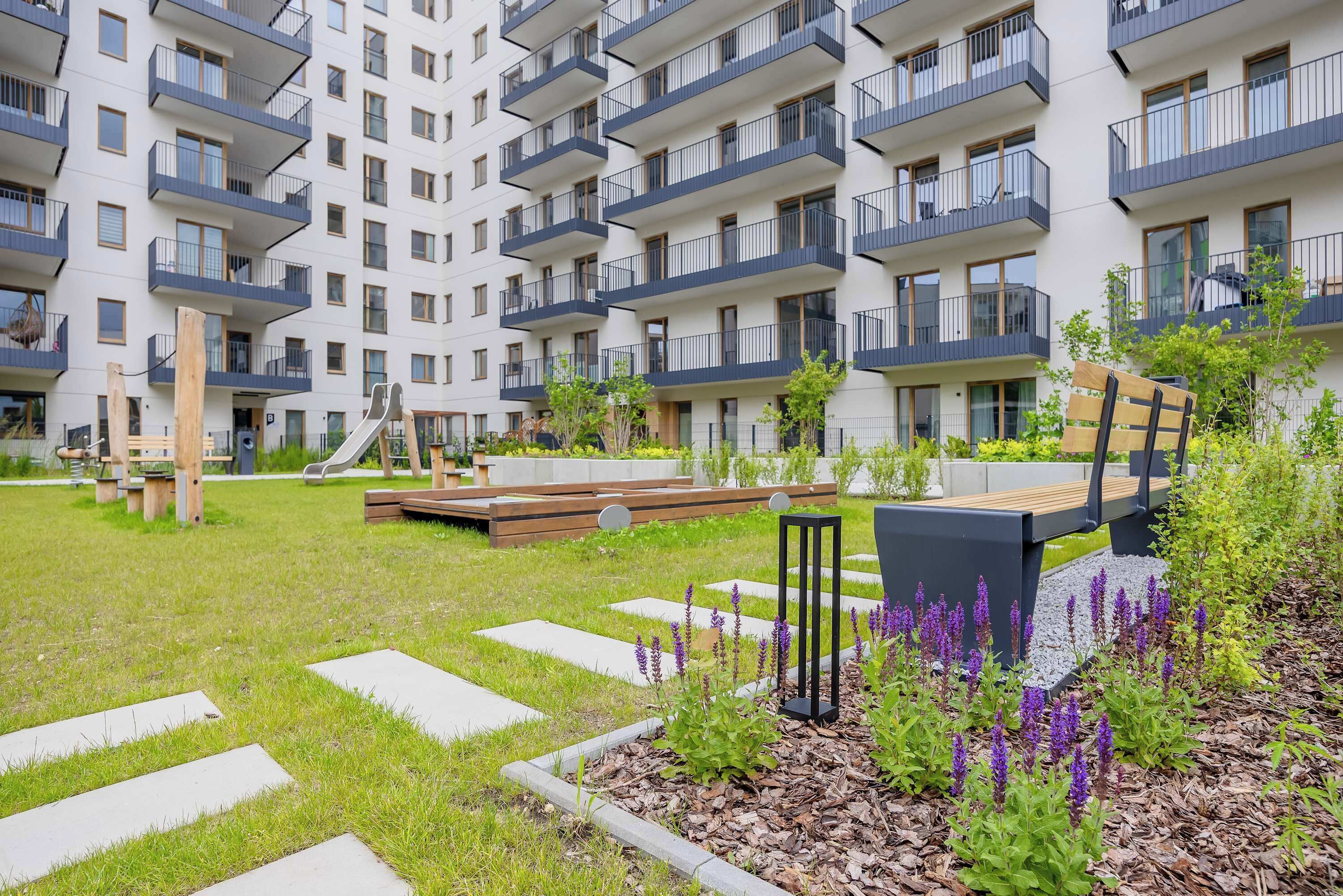Un immeuble résidentiel doté de nombreux balcons, d’une aire de jeux et d’un jardin agrémenté de pas japonais et de plantes.