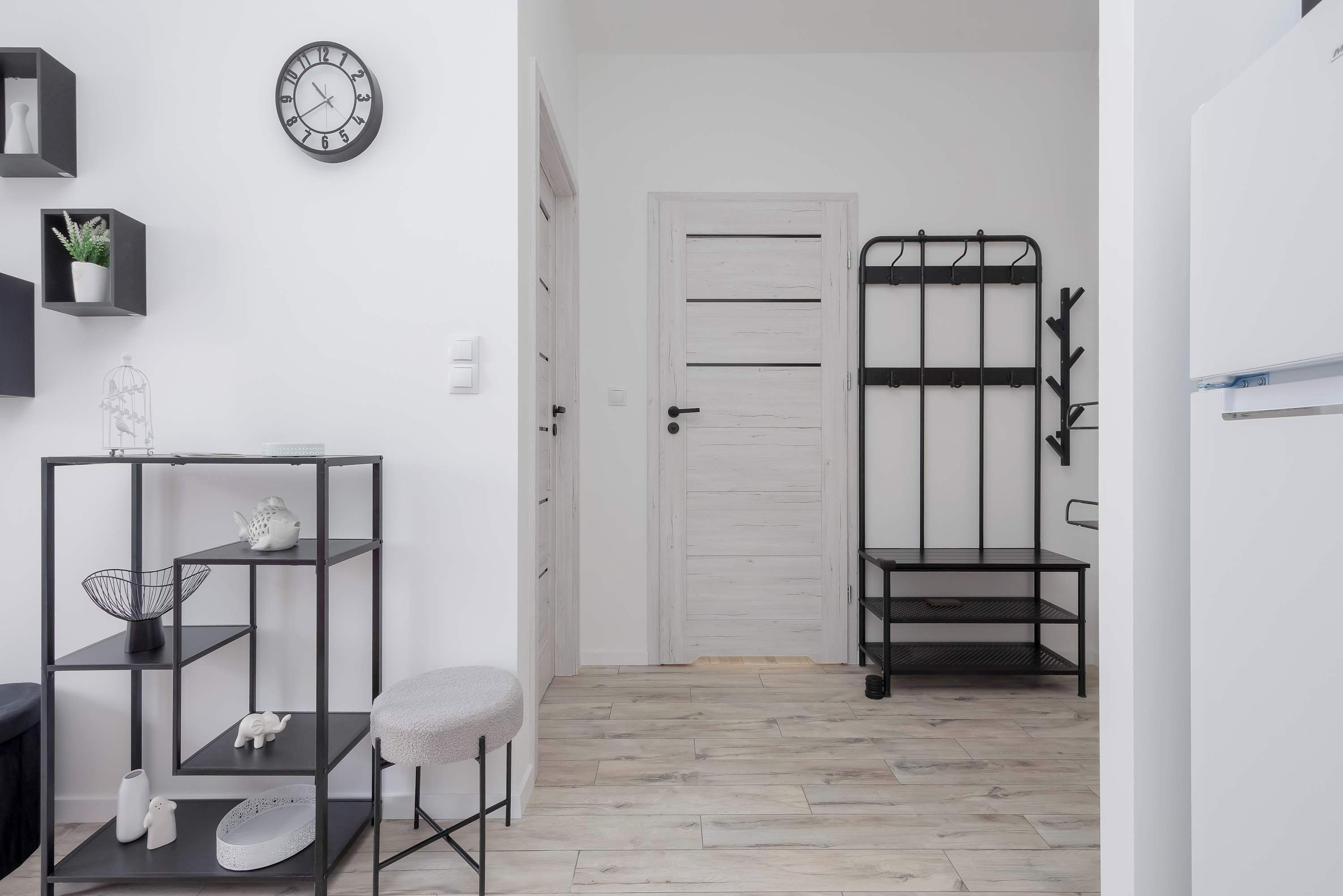 A hallway with a white door, a black metal shelf unit with decorative items, a clock on the wall, and a patterned floor.