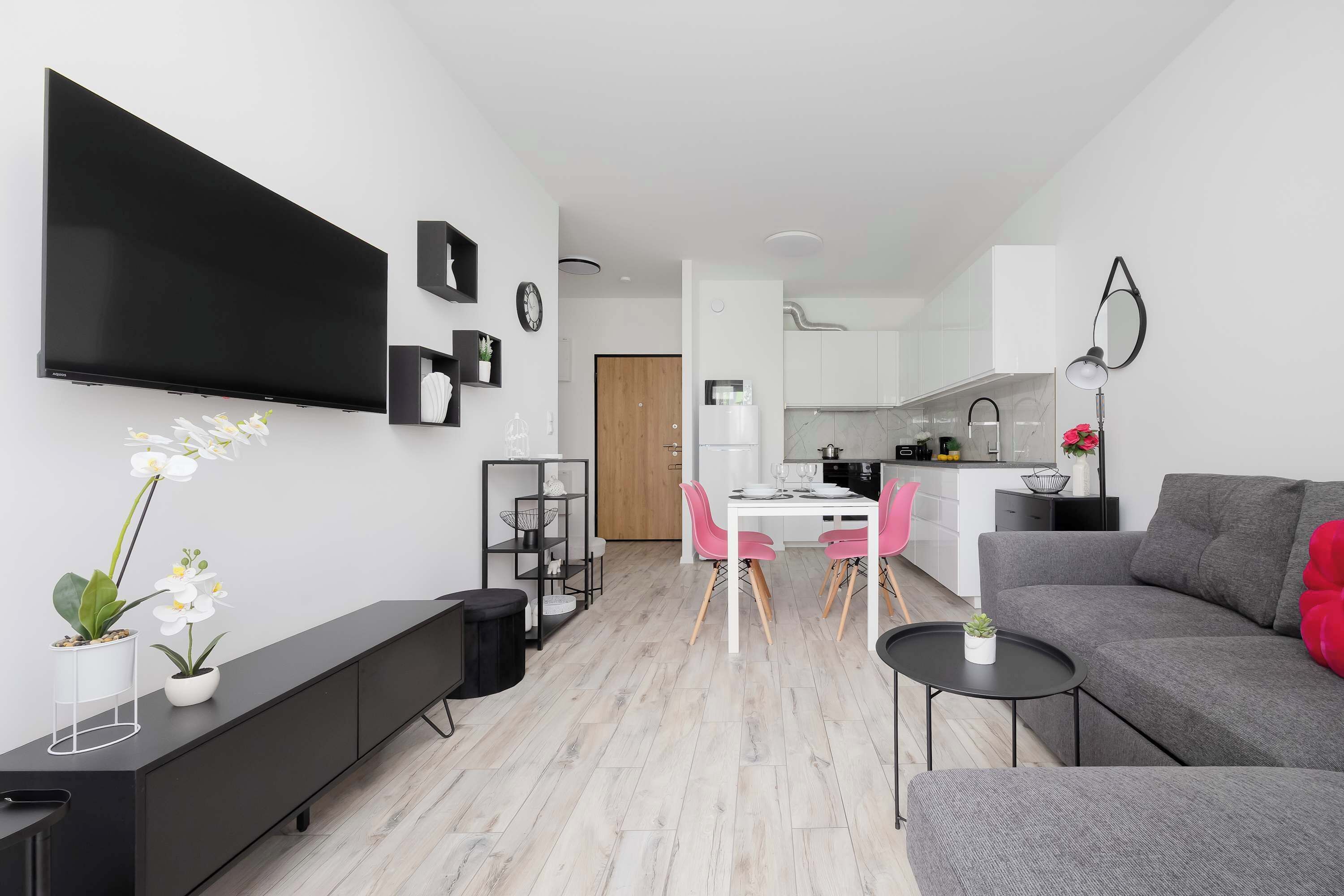 Uma sala moderna com televisão de tela plana, área de jantar com cadeiras cor-de-rosa, sofá cinza e cozinha ao fundo.