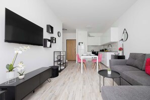 Ein modernes Wohnzimmer mit Flachbildfernseher, einer Essecke mit rosa Stühlen, einem grauen Sofa und einer Küche im Hintergrund.