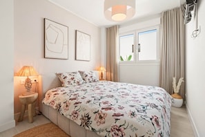 Ein Schlafzimmer mit einem Bett, zwei Nachttischlampen, einem Fenster mit Vorhängen und zwei gerahmten Kunstwerken an der Wand.