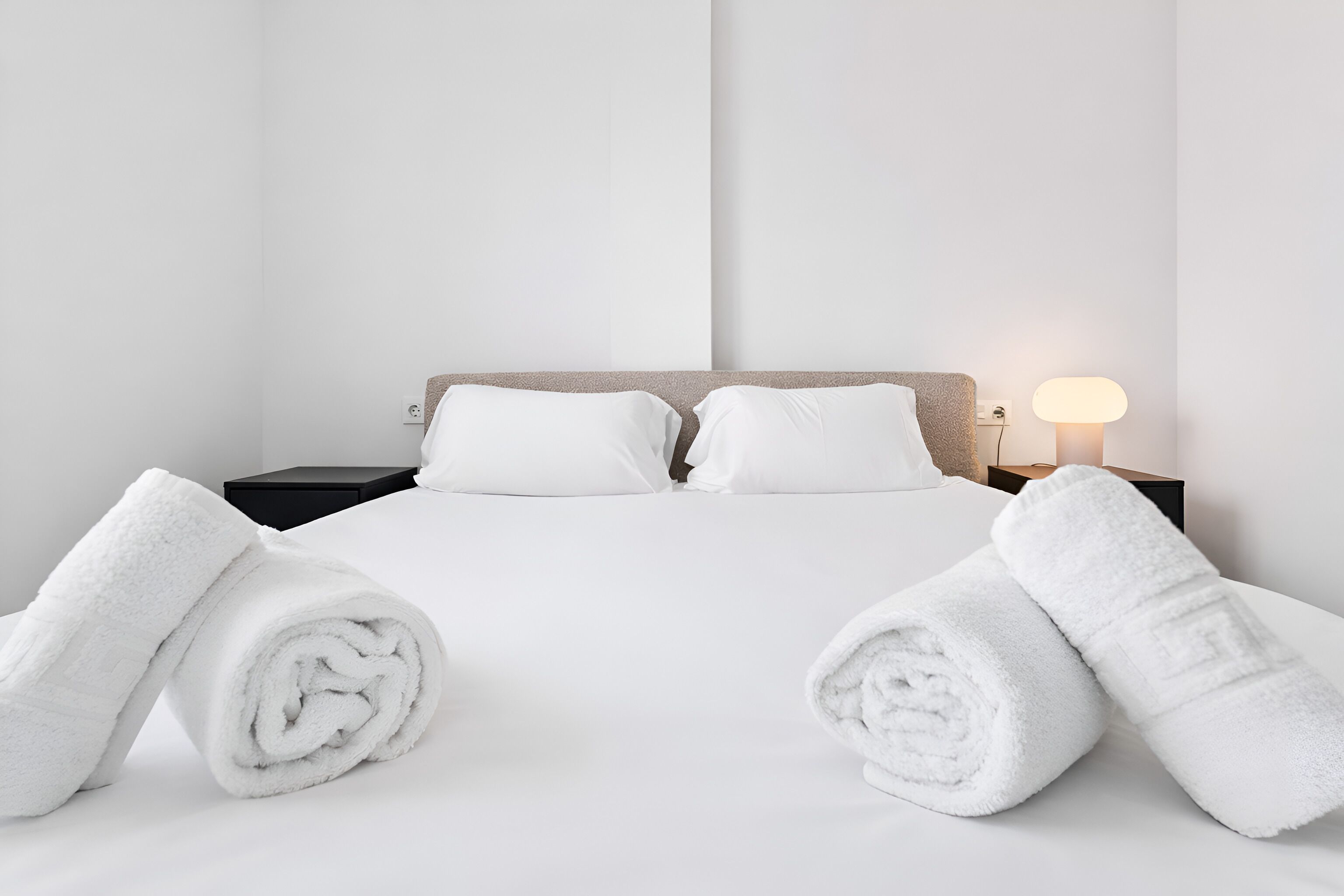 Een hotelkamer met een bed, twee kussens, een nachtkastje, een lamp en twee opgerolde handdoeken.