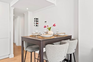 Ein Essbereich mit einem dunklen Holztisch, grau gepolsterten Stühlen, einer Blumenvase und einem Weinkühlschrank an der Wand.