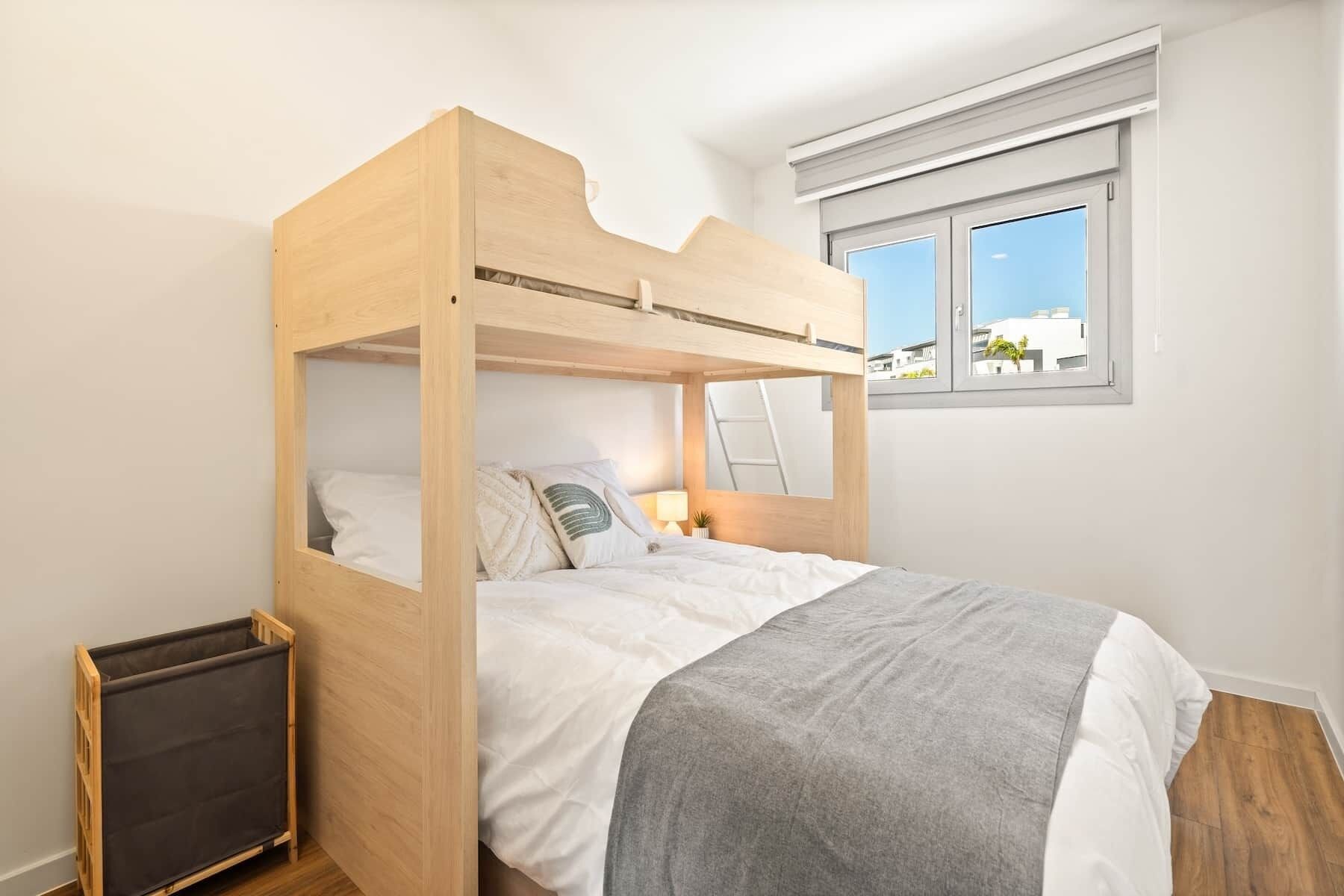 A bunk bed with a wooden frame, white bedding, a grey throw blanket, a small bedside table with a lamp, and a window with a view of buildings.