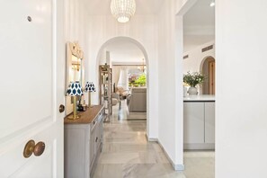 A hallway with a marble floor, white walls, and a chandelier. There is a kitchen area with a white countertop and a vase of flowers. - RDR548 - Stylish 2 bed apt in La Ola (Marbella)