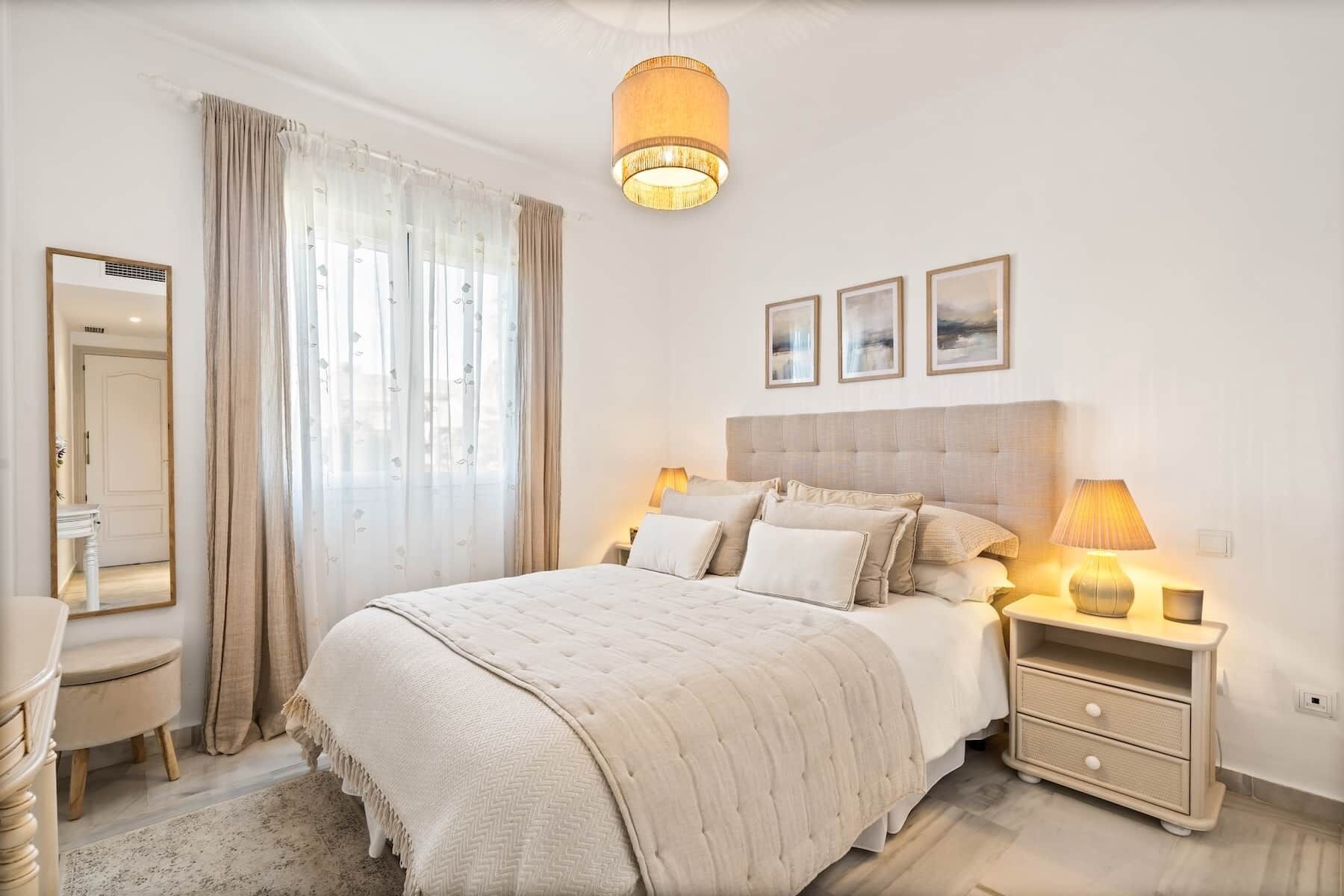 Un dormitorio con cama, mesitas de noche, un espejo y una ventana con cortinas.