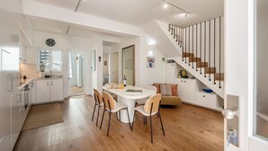 Een moderne keuken met witte kasten, een eethoek met een ronde tafel en stoelen, een trap met zwarte leuningen en een woonruimte met een bank en kussens.
