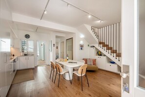 Eine moderne Küche mit weißen Schränken, ein Essbereich mit rundem Tisch und Stühlen, eine Treppe mit schwarzen Geländern und ein Wohnbereich mit Sofa und Kissen.