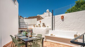 Een terras buiten met een eettafel voor vier personen, omringd door groene stoelen, een barbecue en een witte muur met een decoratieve lantaarn.