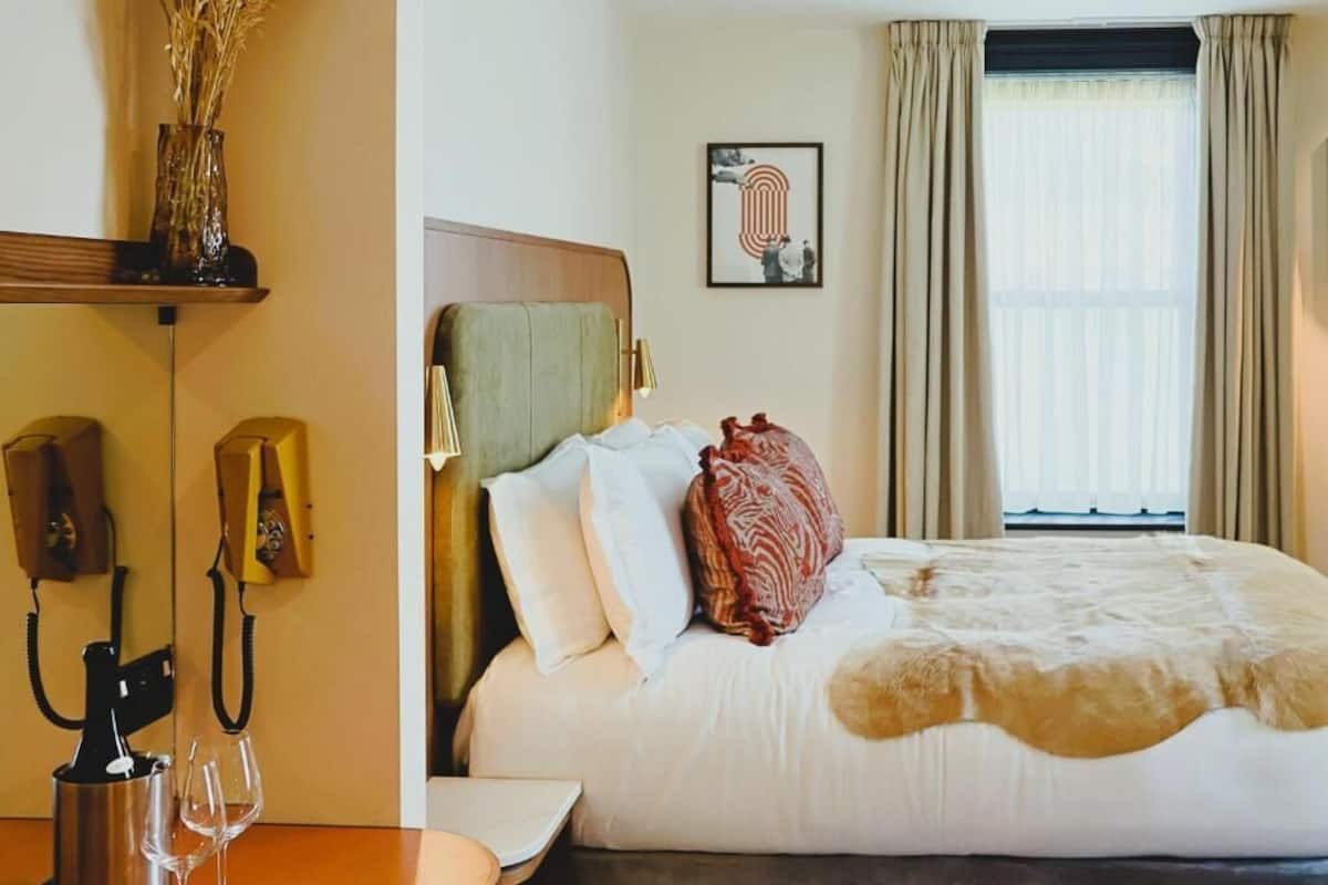 Una habitación de hotel con cama, mesita de noche, lámpara, teléfono y ventana con cortinas.